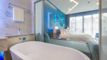 A modern bathroom with a freestanding bathtub, a glass shower enclosure, and a large mirror reflecting a bedroom with a bed and a nightstand.