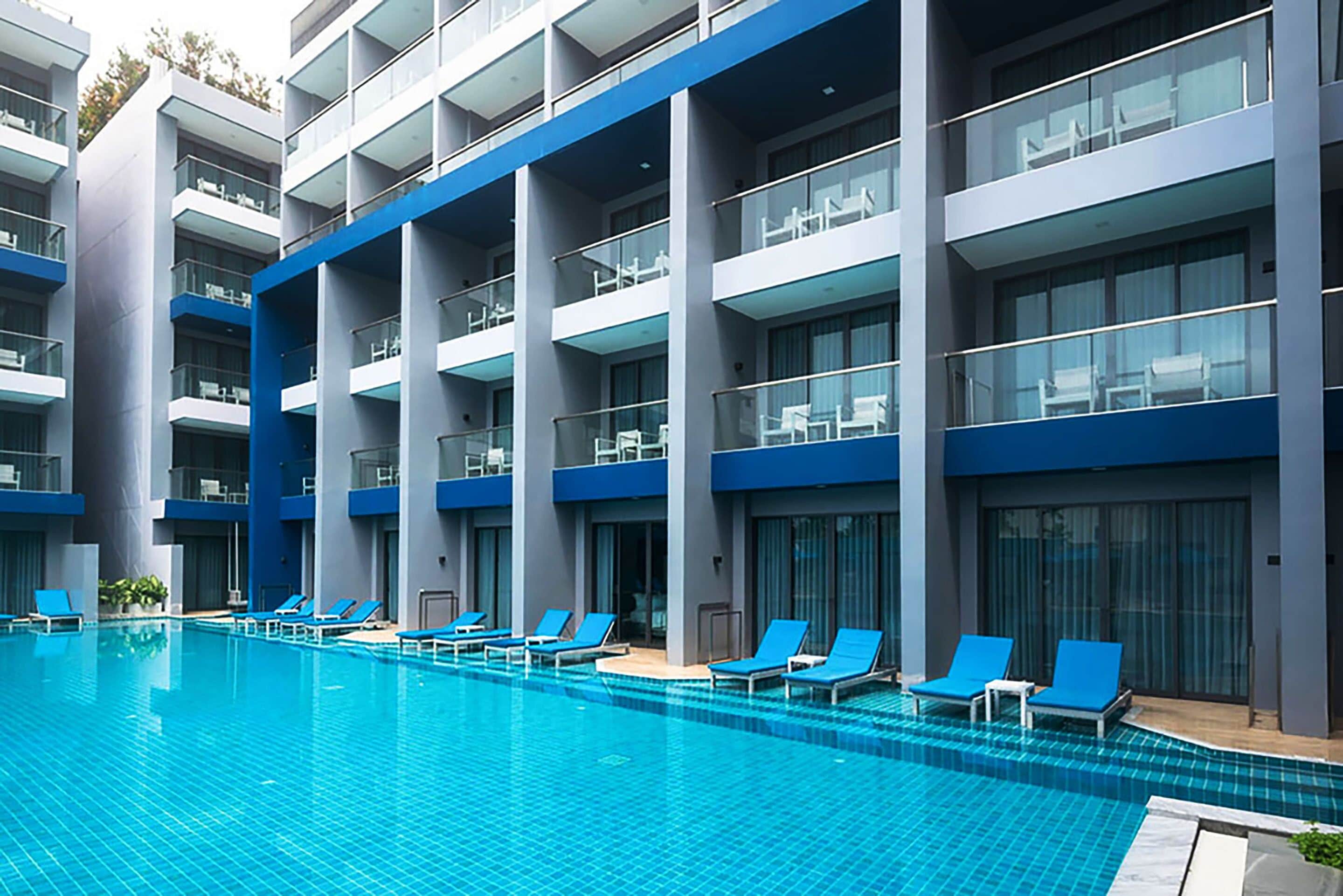 A modern hotel with a large swimming pool, multiple balconies, and blue lounge chairs.