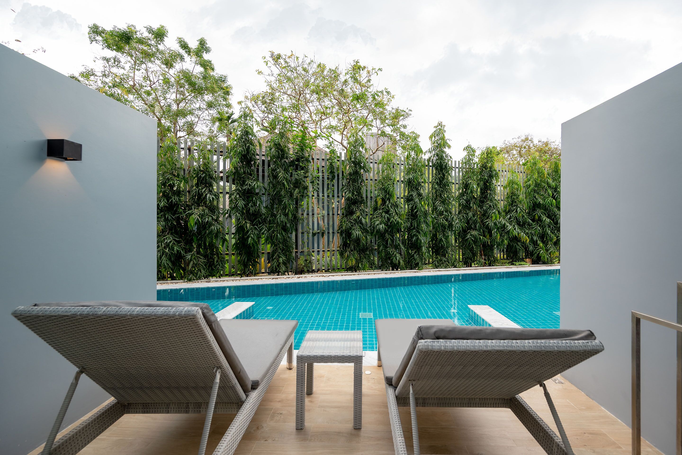 A modern outdoor pool area with two lounge chairs and a small table, surrounded by lush greenery and a wooden fence.