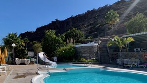 A swimming pool with a slide, surrounded by lounge chairs and umbrellas, with a mountain backdrop. - Ocean view apartment in Playa del Cura Gran Canaria (Mogán)
