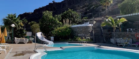 A swimming pool with a slide, surrounded by lounge chairs and umbrellas, with a mountain backdrop.
