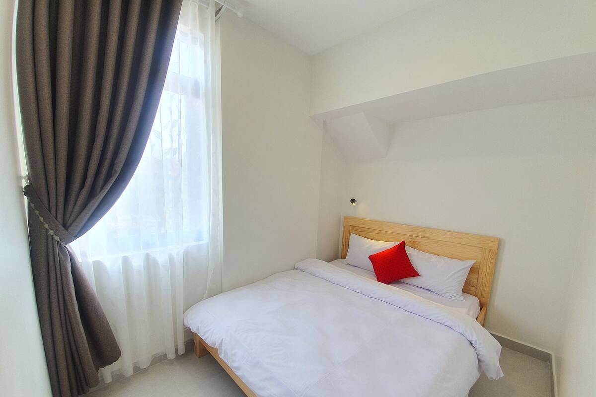 A neatly made bed with white bedding and a red pillow, a wooden headboard, and a window with sheer curtains.