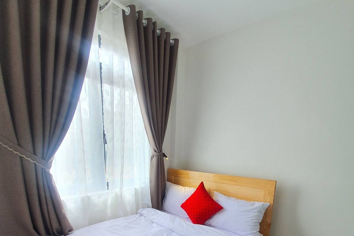 A neatly made bed with white linens and a wooden headboard, a red pillow, and brown curtains partially drawn to the side.