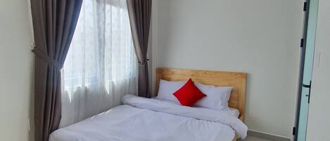 A neatly made bed with white bedding and a red pillow, a wooden headboard, and a window with curtains.