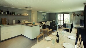 Una cocina moderna con armarios blancos, un comedor con una mesa dispuesta para cuatro personas y una sala de estar con sofá y sillones.