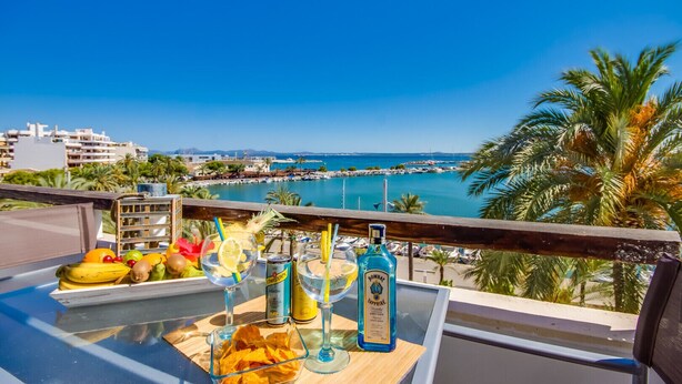 A balcony with a table set for drinks, including bottles and glasses, overlooking a coastal area with buildings and a marina. - Apartment in Alcudia Enjoy near the beach (Alcúdia)