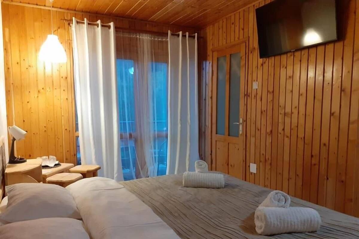 A wooden-paneled room with a bed, a TV on the wall, and a window with curtains.