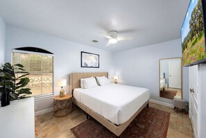 Ein Schlafzimmer mit Bett, Deckenventilator, Fenster mit Jalousien, Nachttisch mit Lampe, Spiegel und Blick auf einen Garten.
