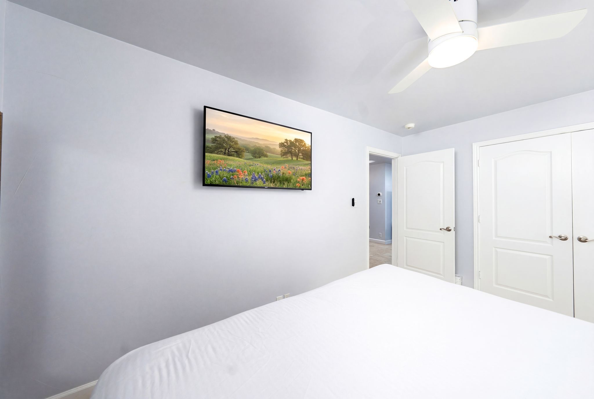 Een slaapkamer met een bed, een plafondventilator, een aan de muur bevestigde televisie en witte deuren.