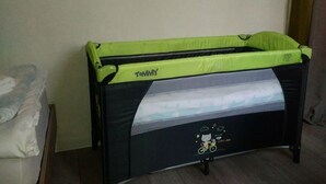 Una culla per neonati nera e verde con una zanzariera, con l'etichetta "Tommy", in una stanza con pavimento in legno e parete neutra.