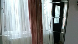 Una stanza con un grande specchio che riflette una finestra con tende bianche trasparenti e una tenda rosa. Il pavimento è in legno e si nota una presa elettrica.