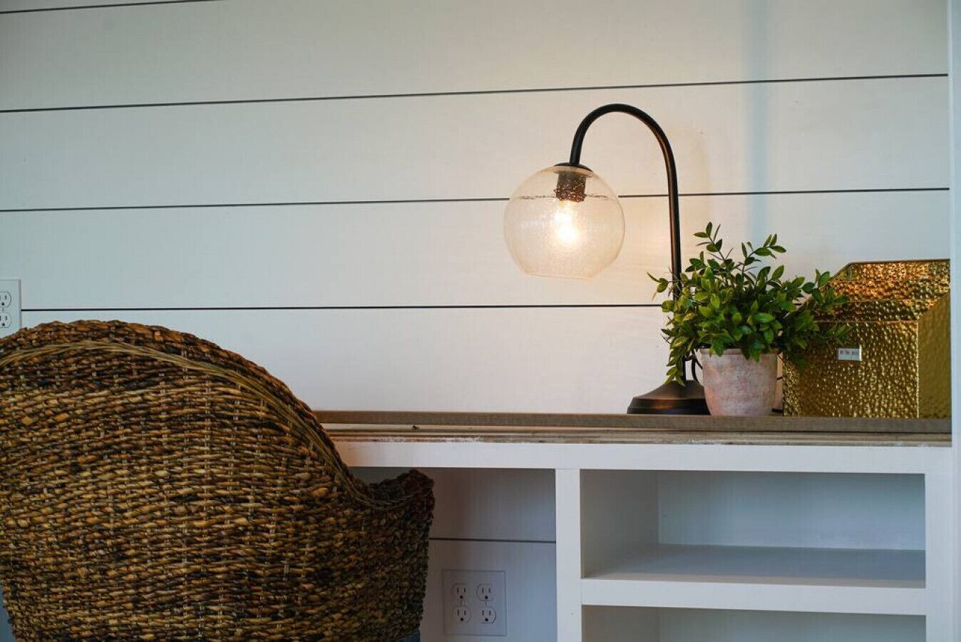 Une chaise en osier, une lampe fixée au mur, une plante en pot et une étagère avec une boîte décorative.