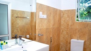 Un baño con lavamanos blanco, espejo y retrete. Hay un perchero sobre el lavamanos y una ventana con vistas a la vegetación.