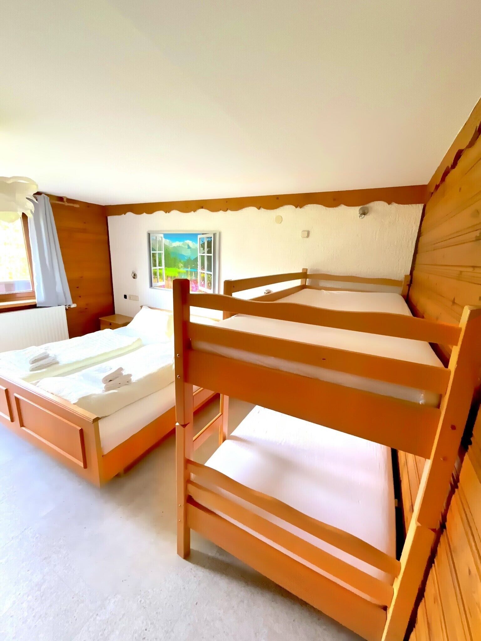 A room with two wooden bunk beds, a window with a view of greenery, and a door with a painting.