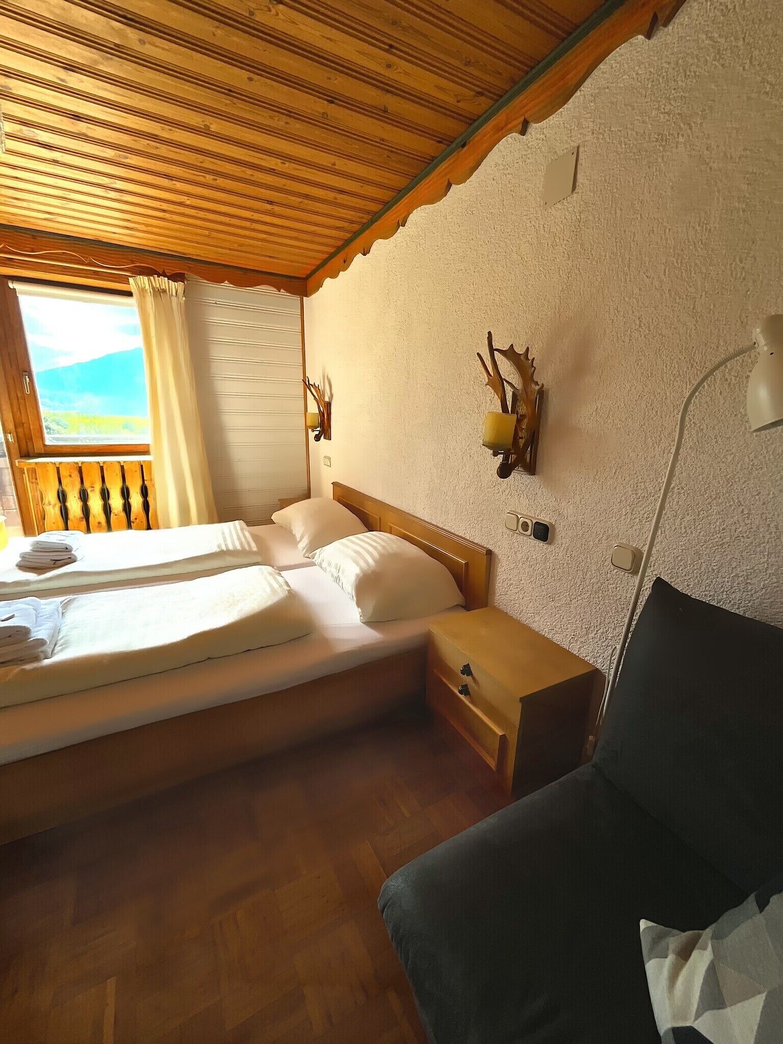 A bedroom with a wooden ceiling, a bed with white linens, a nightstand, a lamp, and a view of the outdoors through a window.