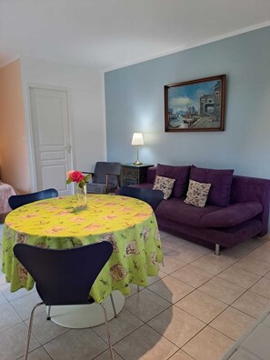 A living room with a round table covered in a yellow tablecloth, a purple sofa, a painting on the wall, and a lamp on a side table. - In the bird paradise, an oasis of silence, the best recreation for you! (Narbonne)