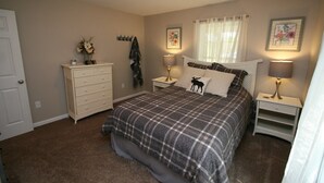 Une chambre à coucher avec une couvre-lit à carreaux, une commode, deux tables de chevet avec des lampes, une fenêtre avec des rideaux légers et une porte.
