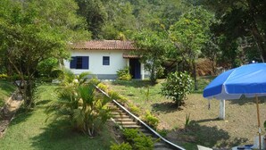 A house with a tiled roof, a small porch, and a garden with a pathway leading to the house. There is a blue umbrella and a white plastic chair near a table.