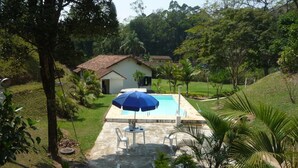 A swimming pool with a blue umbrella, surrounded by greenery and a small white building in the background.
