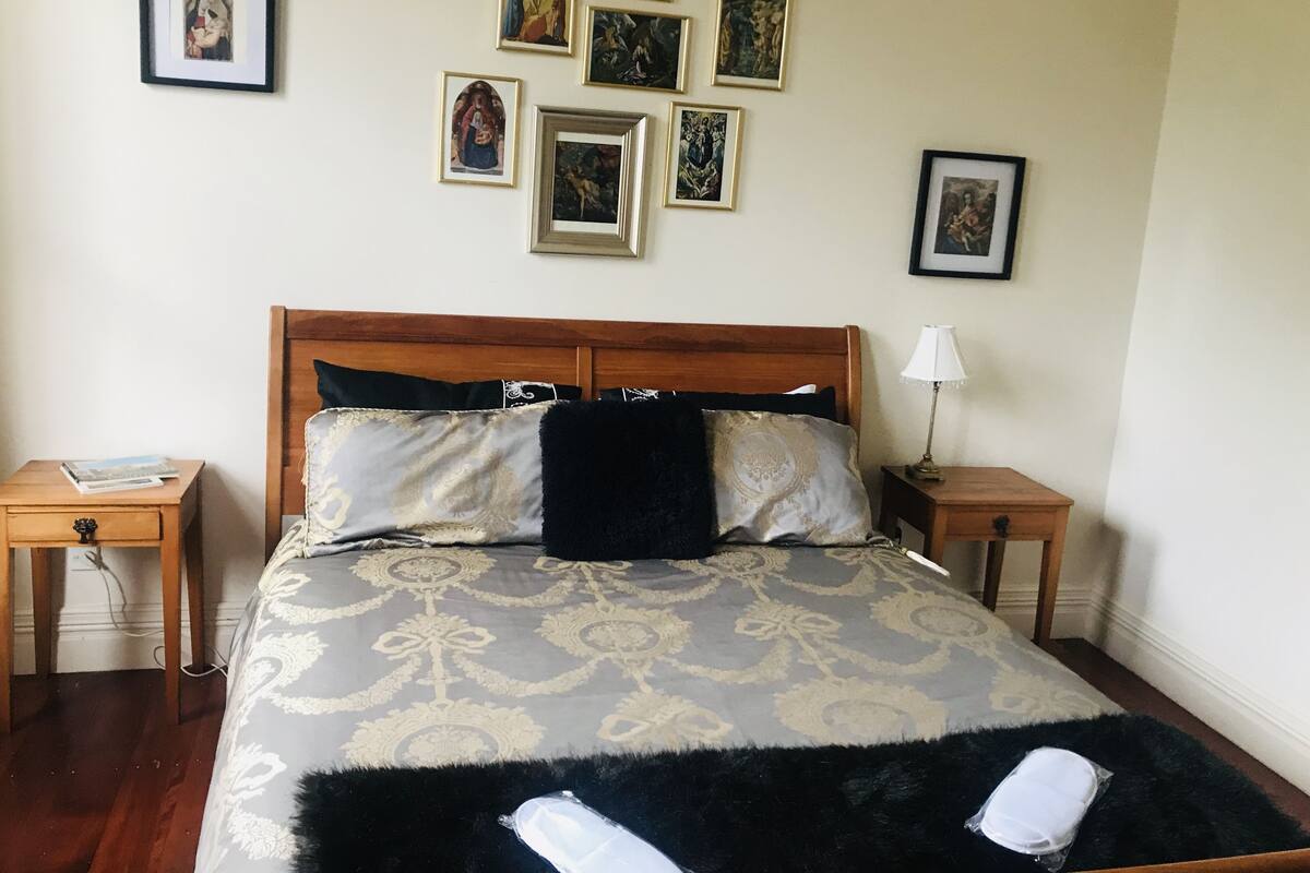 Un dormitorio ordenado con cabecera de madera, colcha estampada y varias fotografías enmarcadas en la pared.