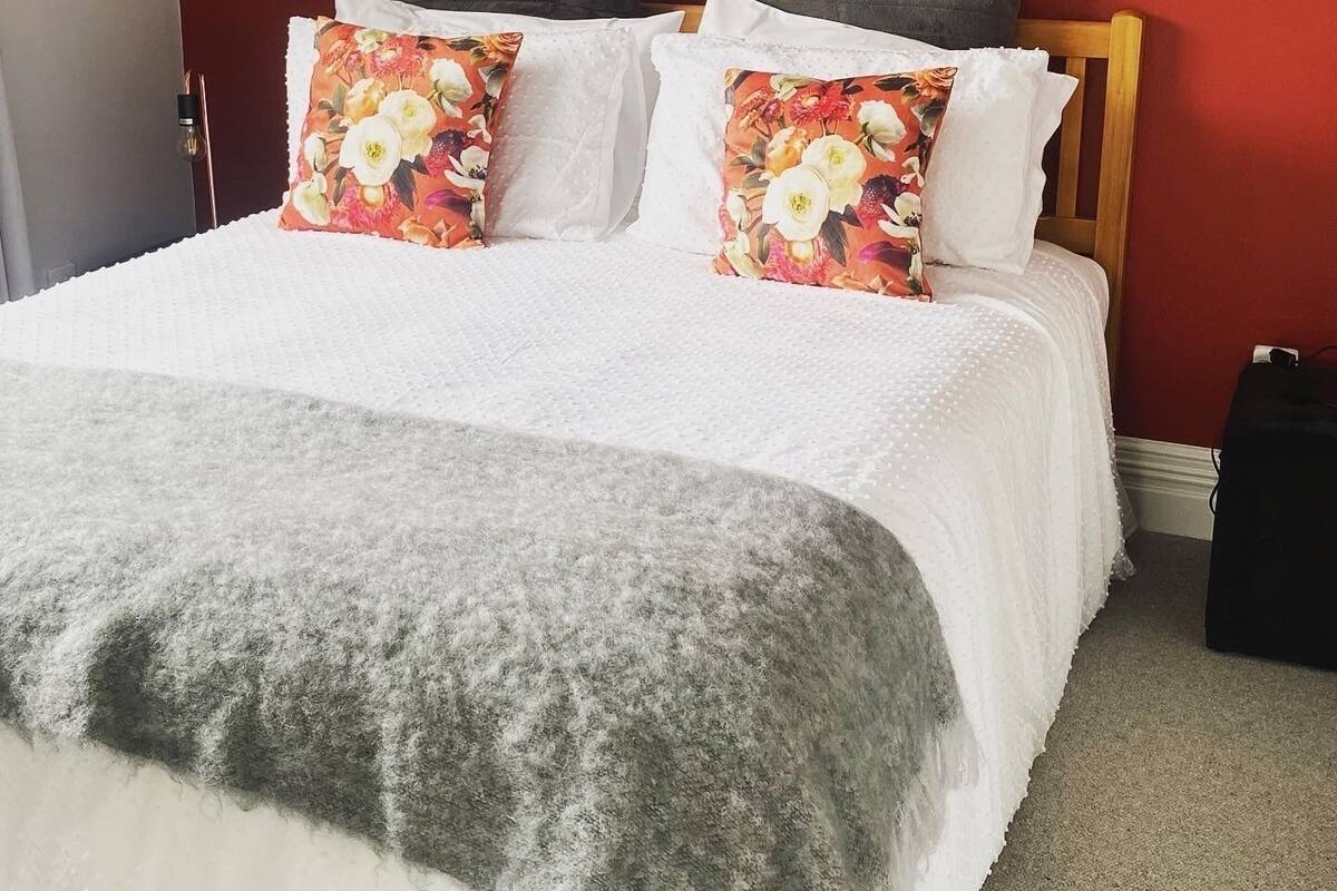 Una cama con ropa de cama blanca, almohadas estampadas con motivos florales, un plaid gris y un cabecero de madera.