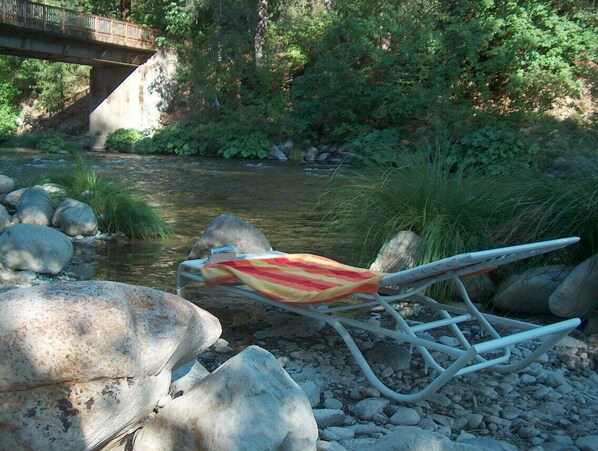 Una silla con un cojín a rayas junto a un río, rocas y un puente al fondo.