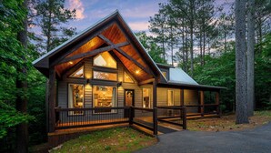 A log cabin with a large deck and multiple windows, surrounded by a forest.