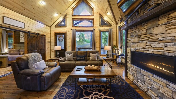 A log cabin interior with a stone fireplace, leather furniture, and large windows overlooking a forested area.