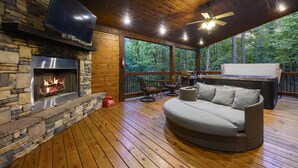 A cozy outdoor deck with a stone fireplace, a flat-screen TV, wicker furniture, and a grill.
