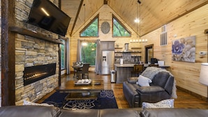 A cozy cabin interior with a stone fireplace, a dining area, a kitchen, and a living room with a leather couch and a large window.
