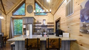 A wooden cabin interior with a kitchen island, bar stools, and a large window overlooking greenery.