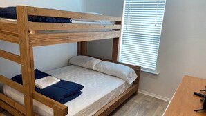 Un lit superposé avec une structure en bois, un lit inférieur avec une literie blanche et des oreillers bleus, et une fenêtre avec des stores.
