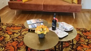 Un salon avec un canapé marron, deux petites tables rondes, un vase avec des fleurs, une bouteille de vin et quelques magazines.