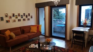 Un salon avec un canapé marron, une petite table ronde sur laquelle se trouvent un magazine, une bouteille et un vase rempli de fleurs. Il y a un balcon avec une table et des chaises, et un lustre au-dessus.