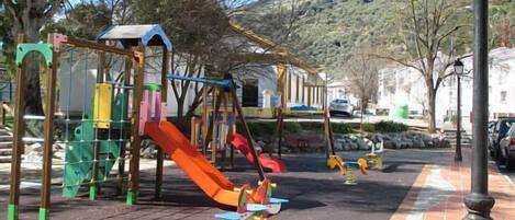 A playground with swings and slides, surrounded by trees and a hilly landscape.
