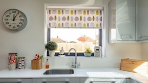 Una cocina con un lavamanos, un reloj, una ventana con persiana de motivos de hojas y vista a un patio trasero.