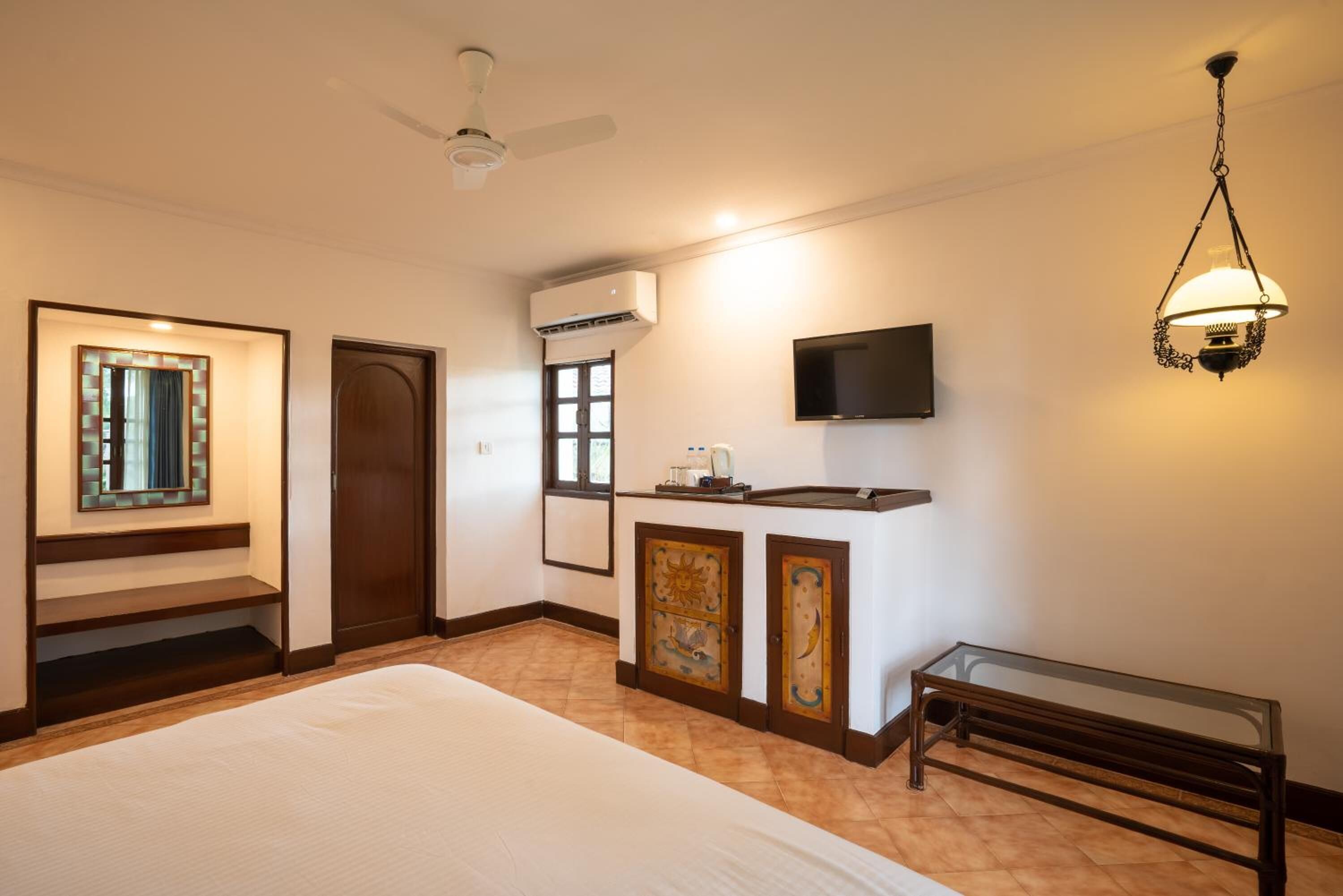 Quarto de hotel com cama, escrivaninha com TV de tela plana, ventilador de teto, ar condicionado e luminária decorativa.
