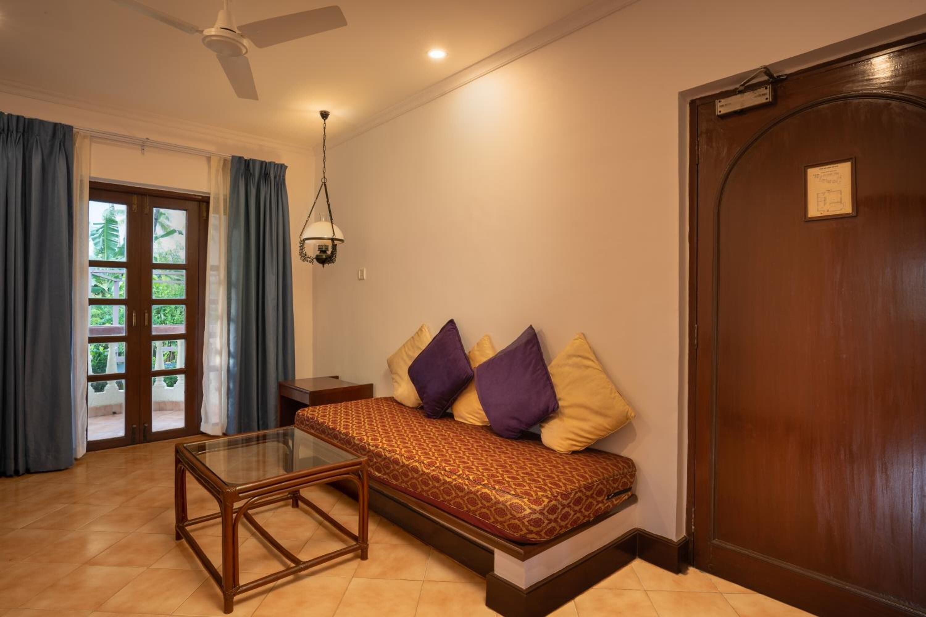 A room with a sofa bed, a glass coffee table, a ceiling fan, and a door with a plaque.