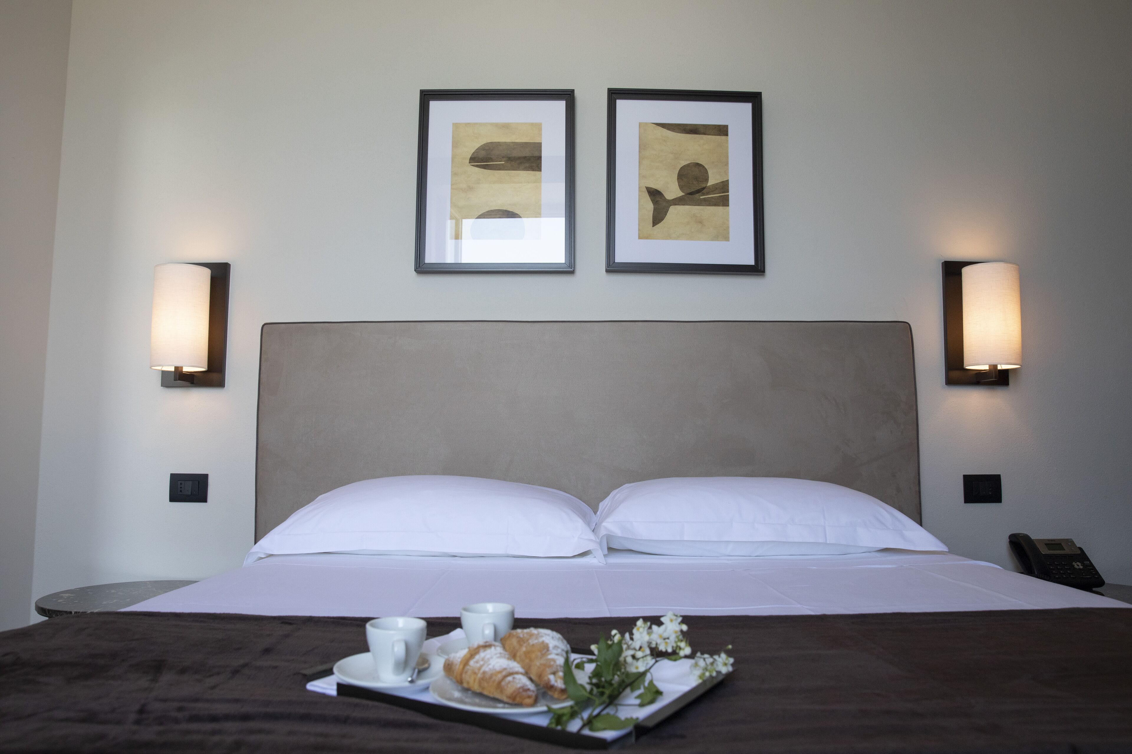 Habitación de hotel con una cama, dos cuadros en la pared, lámparas de noche, una bandeja con croissants y una taza, y un teléfono en la mesita de noche.