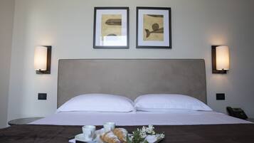 Habitación de hotel con una cama, dos cuadros en la pared, lámparas de noche, una bandeja con croissants y una taza, y un teléfono en la mesita de noche.
