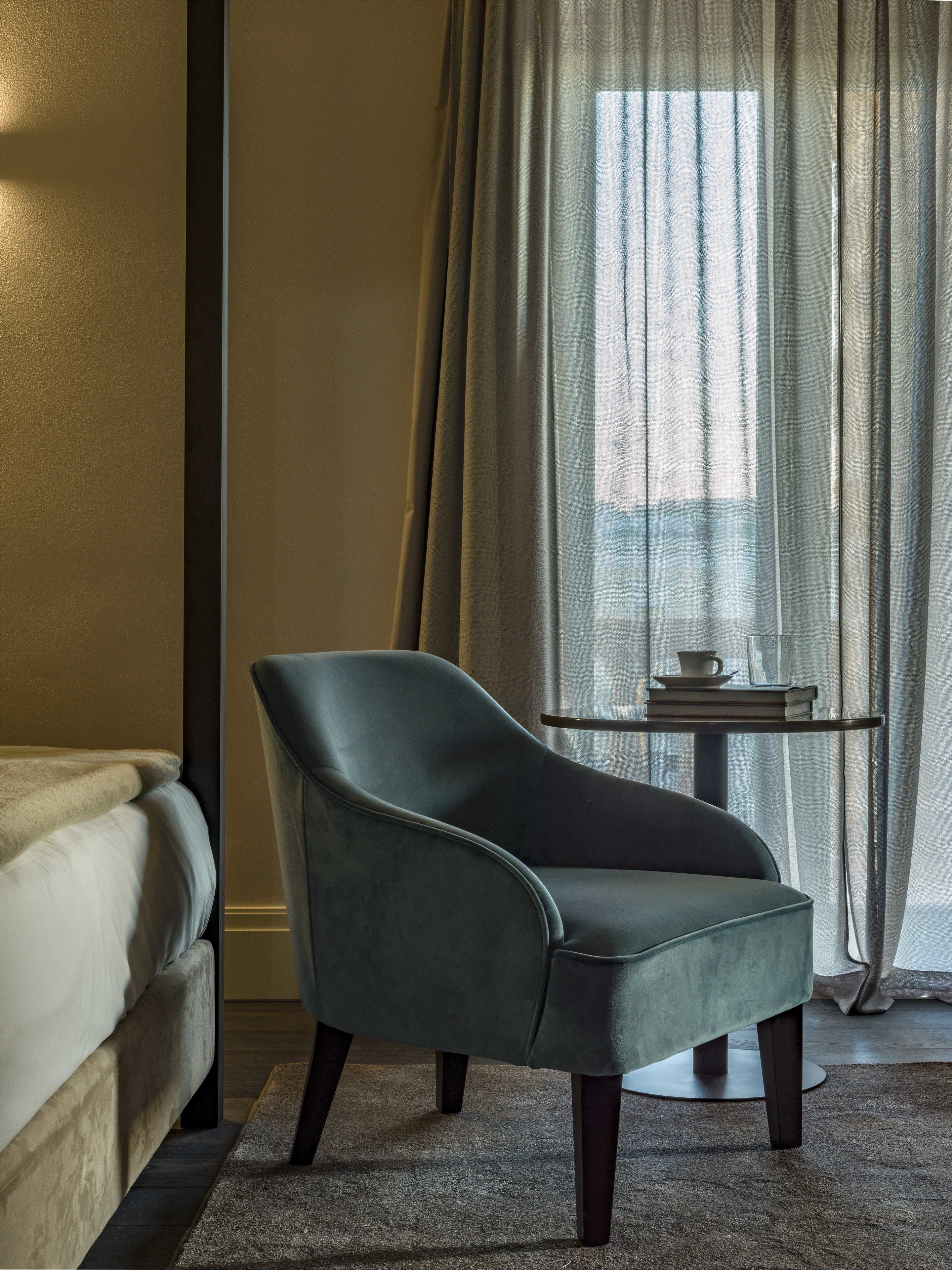 A modern room with a grey armchair, a small round table with a cup and book, and a bed with a white headboard.