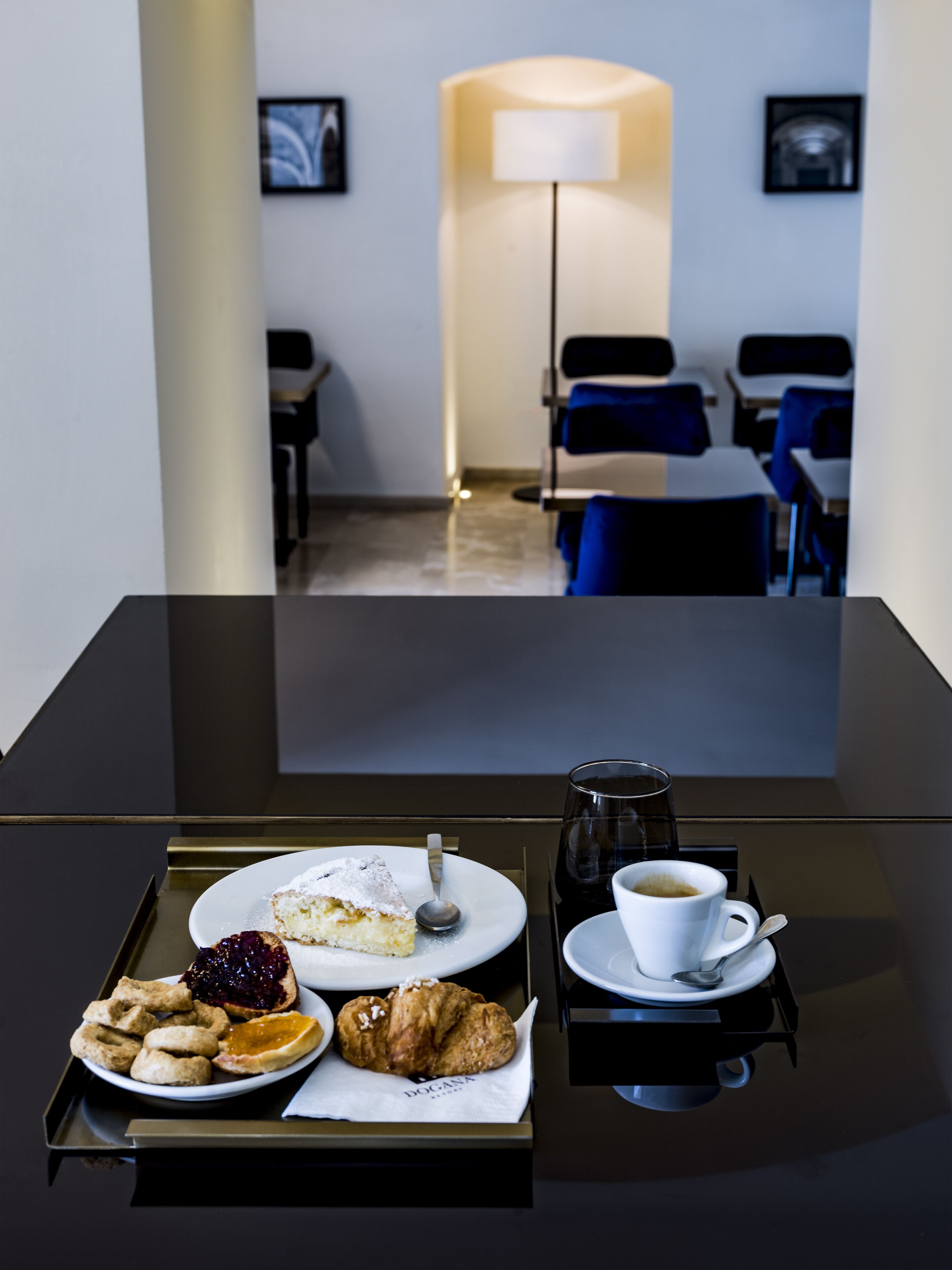 A hotel room with a dining table set with a slice of cake, a cup of coffee, and various pastries. There is a lamp and a bed in the background.