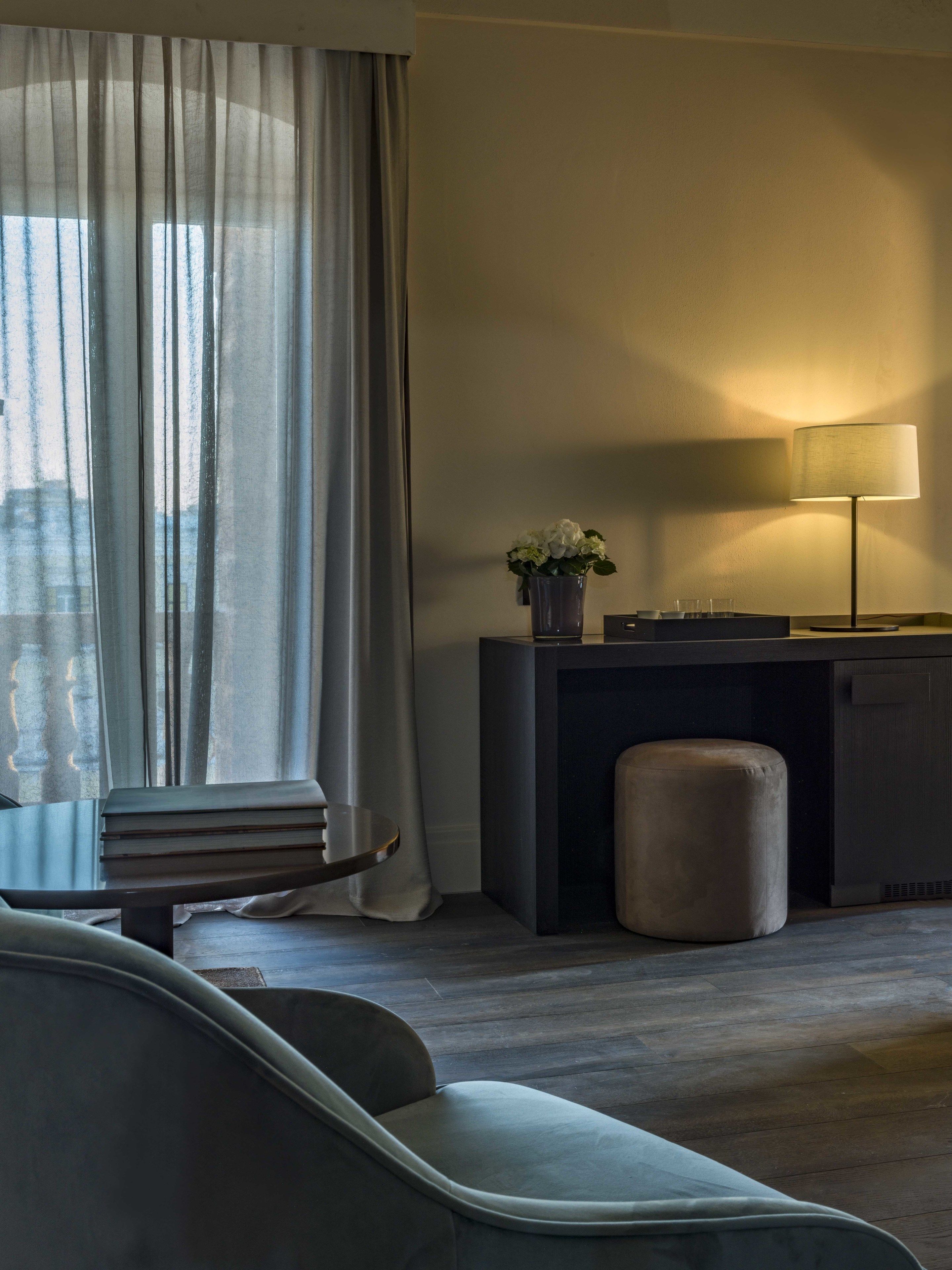 A modern hotel room with a sofa, a desk with a lamp, a vase with flowers, and a view through sheer curtains.