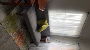 A room with a grey sofa, a yellow pillow, a patterned rug, and a window with sheer curtains.