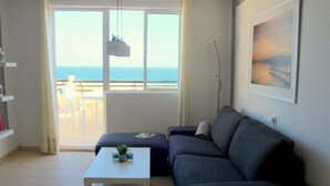 A living room with a sofa, a coffee table, and a view of the sea through a large window.