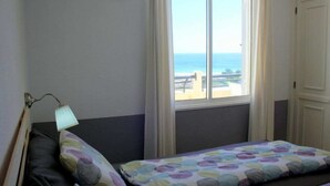 A bedroom with a bed, a nightstand, a lamp, a window with a view of the sea, and a door.