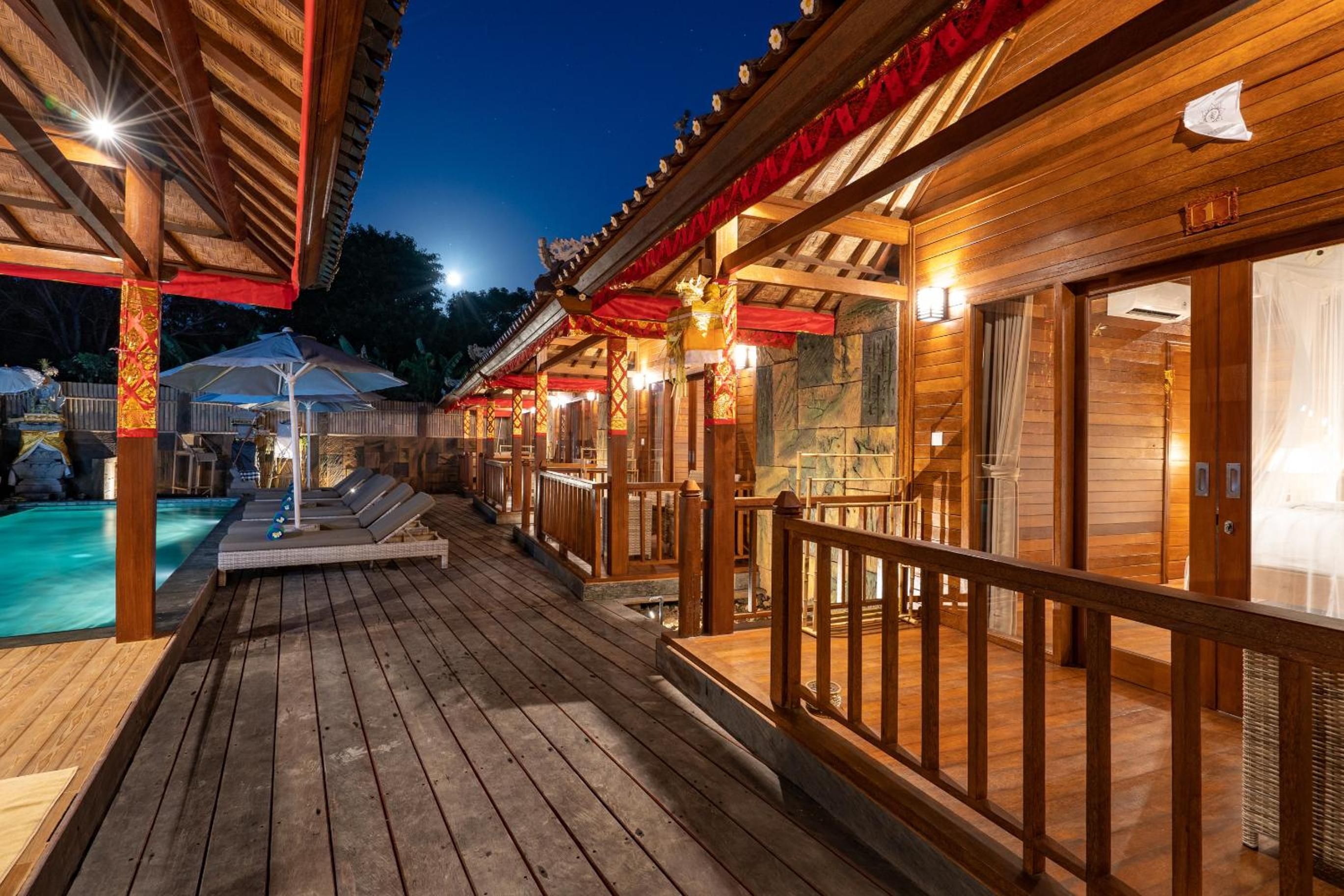 Una terraza de madera con piscina, edificios de madera con toldos rojos y un cielo nocturno.