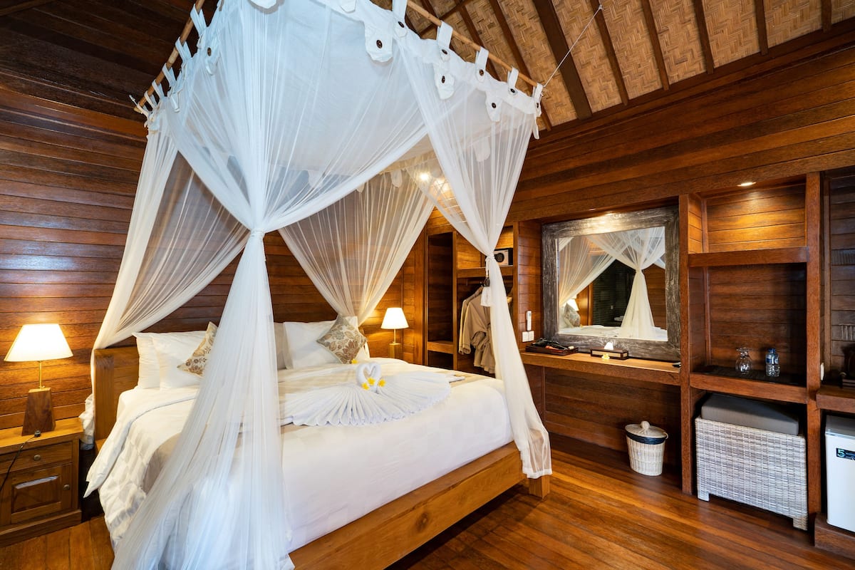 Een hemelbed met witte lakens, een houten hoofdbord, nachtlampjes en een klein nachtkastje.