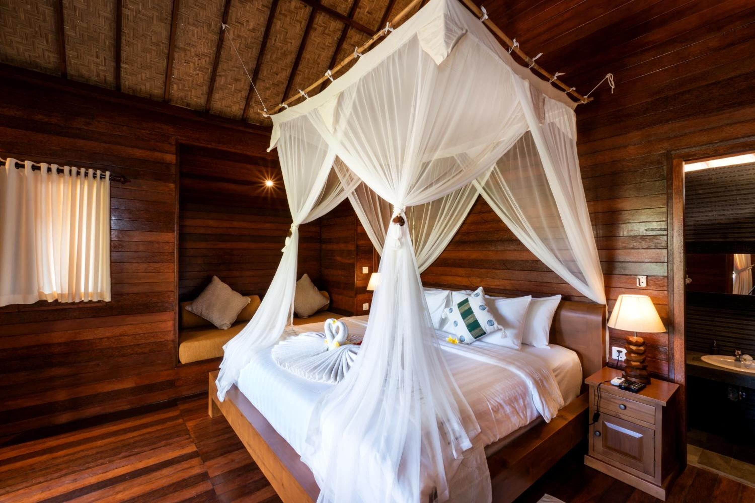 Ein Zimmer mit Holzboden, einem Himmelbett, einem Nachttisch mit Lampe und einem mit Vorhängen drapierten Fenster.