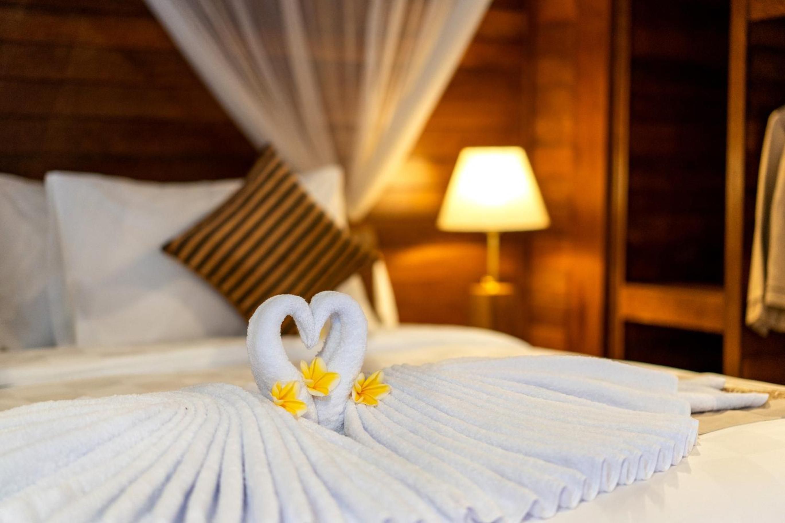 A hotel room with a neatly made bed, a heart-shaped towel arrangement, and a lamp on the side.